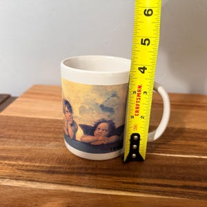 Op de afbeelding: Een witte keramische mok met een afbeelding van twee cherubijntjes tegen een blauwe lucht. Een geel Craftsman-meetlint is verticaal tegen de zijkant van de mok geplaatst en geeft een hoogte van ongeveer 9,5 cm aan.
