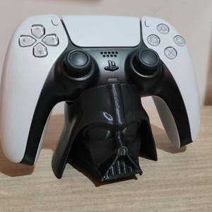 Könnte beinhalten: Ein weißer PlayStation 5-Controller liegt auf einem schwarzen Darth-Vader-Themenständer. Der Controller hat schwarze Akzente und Knöpfe. Der Ständer ist eine detaillierte Nachbildung von Darth Vaders Helm und verleiht dem Gaming-Setup eine einzigartige Note.