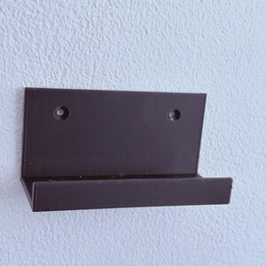 May include: A dark brown, L-shaped wall-mounted shelf featuring a flat back panel with two circular pre-drilled holes for mounting and a horizontal ledge extending outward to support items. The surface has a visible, fine-textured finish.