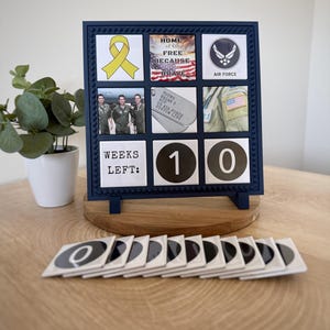 May include: A square navy blue frame with a grid of nine tiles featuring a yellow ribbon, an American flag graphic with text, the Air Force logo, a photo of three people, dog tags, a uniform patch, and the text WEEKS LEFT: 1 0.