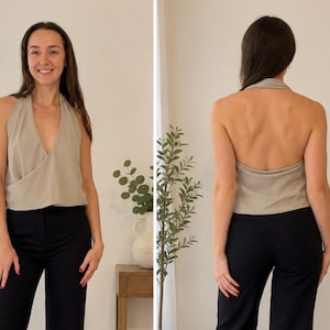 Op de afbeelding: Een beige haltertop met een diepe V-hals, een overslag aan de voorzijde en een open rug. Het kledingstuk is mouwloos en gemaakt van een gladde, effen stof, gecombineerd met een eenvoudige zwarte broek.