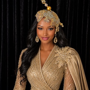 May include: A gold sequined wrap dress with three-quarter sleeves and a matching capelet featuring a large crystal-encrusted floral brooch. The ensemble is paired with a gold headpiece adorned with crystals and beads, and teardrop earrings.