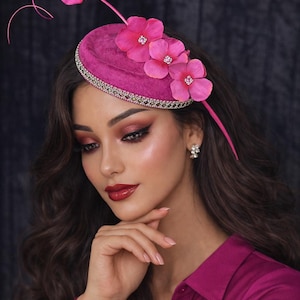 May include: A fuchsia-colored fascinator with a rounded base, adorned with three large, pink fabric flowers and a delicate beaded trim. The headpiece features a thin, curved stem with a single flower and is designed for special occasions.