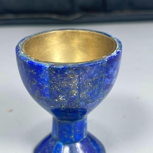 Op de afbeelding: Een kleine kelk met een diepblauwe lapis lazuli buitenkant, bestaande uit meerdere ingelegde steensegmenten met zichtbare goudkleurige spikkels. De binnenkant van de beker is bekleed met een gladde, gepolijste messingkleurige metalen afwerking.