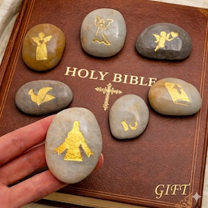 May include: A collection of smooth, oval-shaped stones with gold religious symbols, including angels, a dove, and praying hands. The stones are arranged on a brown leather-bound Holy Bible, with the word "GIFT" visible.