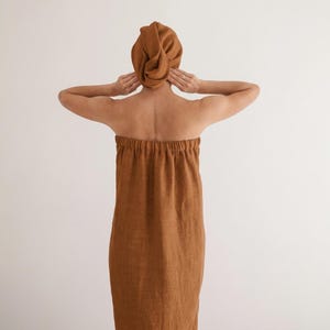 Puede incluir: Vista trasera de un conjunto de spa de lino marrón que consta de una toalla de baño y una toalla para el cabello. La toalla de baño es un diseño tubular simple con una parte superior elástica. La toalla para el cabello está envuelta en forma de turbante.