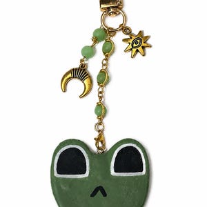 May include: A gold-toned keyring featuring a green frog face charm with large black eyes and a small mouth. The frog is attached to a gold chain with light green beads, a gold crescent moon charm, and a gold star charm with an eye symbol.