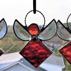May include: Three stained glass angels hanging in a window. Each angel is red and white with a halo. The angels are made of glass and metal.