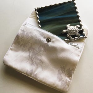 May include: A white fabric pouch with a zipper closure and a silver button. The pouch is decorated with a floral pattern. A small silver framed mirror with a blue glass insert and a silver turtle design is partially visible inside the pouch.