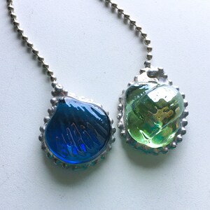 May include: Two glass pendants with silver-colored metal edging and ball chains. One pendant is a deep blue color, and the other is green. The glass has a textured, rippled appearance, and the metal has a decorative, scalloped edge.
