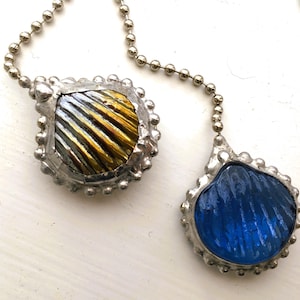 May include: A silver-toned necklace featuring two pendants. One pendant has a square shape with a striped pattern in gold, silver, and black. The other pendant is a blue, shell-shaped design. Both pendants have a textured, beaded border.