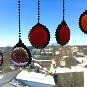 May include: A collection of hanging stained glass suncatchers in various colors, including red, orange, and iridescent hues. Each suncatcher is framed in a black, scalloped metal border and suspended by a beaded chain. The background shows a cityscape.