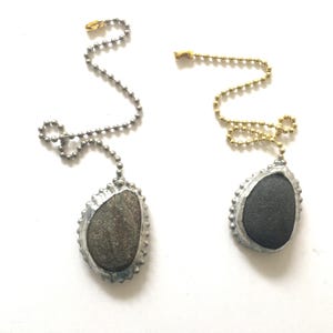 May include: Two handcrafted light pull chains. One features a gray stone set in a silver-colored metal frame, suspended from a silver ball chain. The other has a black stone in a silver frame, hanging from a gold-colored ball chain. Both have a decorative, beaded edge.