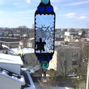May include: A stained glass suncatcher with a silhouette of a sea turtle in the center. The suncatcher is made of clear glass with a textured surface and is surrounded by blue and green glass beads. A crystal prism hangs from the bottom of the suncatcher.