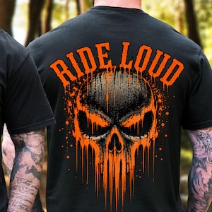 Puede incluir: Una camiseta negra de manga corta con un gráfico en la parte posterior. El diseño muestra una calavera estilizada en tonos negros y naranjas con un efecto de pintura que gotea. Sobre la calavera, el texto RIDE LOUD está impreso en naranja.