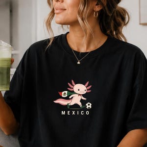 May include: A black short-sleeved t-shirt featuring a graphic of a pink axolotl wearing a cape with the colours of the Mexican flag and kicking a football. The word MEXICO is printed in white capital letters underneath the axolotl illustration.