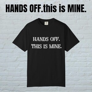 以下が含まれることがあります： 前面に白い文字で「HANDS OFF. THIS IS MINE.」とプリントされた黒いTシャツ。Tシャツは半袖とクルーネックです。「HANDS OFF.this is MINE.」というフレーズがTシャツの上にプリントされています。