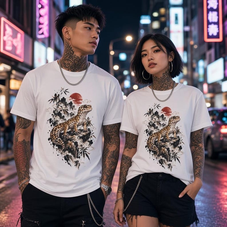 Może przedstawiać: Biały t-shirt z kr&oacute;tkim rękawem z nadrukiem graficznym przedstawiającym gekona lamparciego siedzącego na skale, w otoczeniu łodyg bambusa i czerwonego słońca. Wz&oacute;r wykonany jest w szczeg&oacute;łowym stylu tuszu z czarnymi, białymi i czerwonymi akcentami.