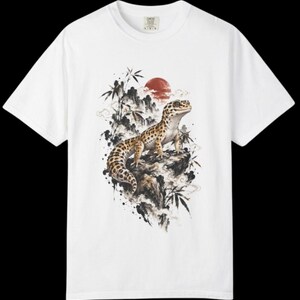 Może przedstawiać: Biały t-shirt z kr&oacute;tkim rękawem z nadrukiem graficznym przedstawiającym cętkowanego gekona lamparciego siedzącego na skale. Wz&oacute;r zawiera czarne pociągnięcia pędzla, łodygi bambusa i czerwone koło słońca w tle, tworząc szczeg&oacute;łową kompozycję.