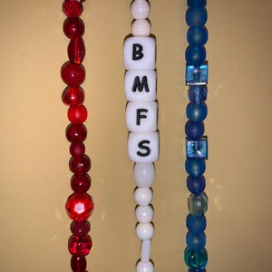 May include: Three beaded strands in red, white, and blue. The red strand features various shapes of red beads. The white strand has lettered beads spelling "BMFS". The blue strand has blue beads and square blue accents.