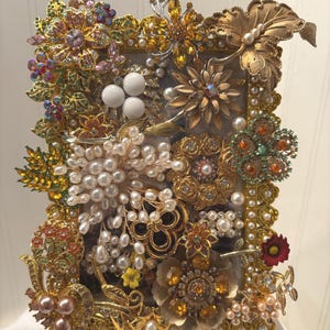 May include: A rectangular shadow box filled with vintage brooches. The brooches are in various shapes and sizes, including flowers, leaves, and abstract designs. The brooches are in gold, silver, and various colors, with pearls and rhinestones. The frame is gold.