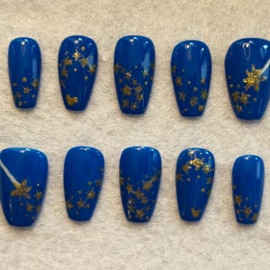 May include: A set of ten artificial nails in a vibrant blue hue, adorned with gold glitter star accents. Some nails feature a white diagonal stripe. The nails are arranged on a white surface, showcasing a celestial theme.