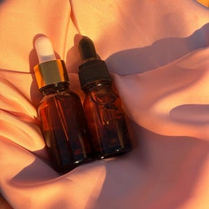 May include: Two small amber glass dropper bottles with gold and black caps are nestled in a peach-colored fabric. The bottles are filled with a clear liquid. The lighting creates shadows and highlights on the fabric and bottles.