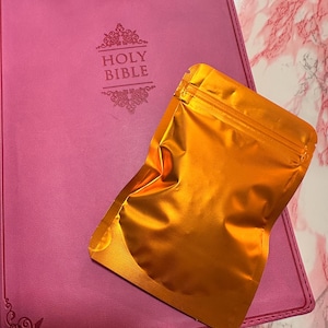 Può includere: Una Bibbia rosa rilegata in pelle con scritte dorate e decorazioni angolari. Una bustina metallica arancione è appoggiata sopra la Bibbia. La copertina della Bibbia presenta le parole "HOLY BIBLE".