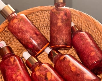 May include: Six clear glass dropper bottles filled with a shimmering, deep red liquid containing visible botanical pieces. Each bottle features a metallic gold collar and a white rubber dropper top, all arranged together inside a woven wicker basket.