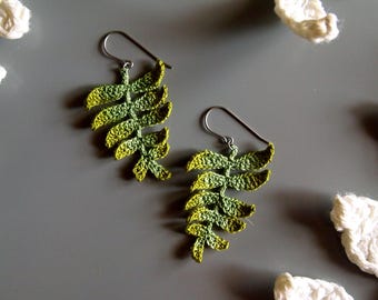 May include: A pair of drop earrings featuring crocheted fern leaf shapes in a gradient of green and yellow thread. The earrings are attached to silver-toned metal hooks and are displayed alongside white crocheted leaf accents.