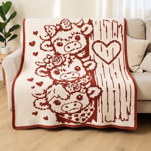 Peut inclure: Une couverture en crochet couleur crème présentant un motif graphique rouge foncé de trois vaches empilées, chacune avec des détails floraux ou à motifs, entourées de petits cœurs et d'une texture de grain de bois verticale avec un grand cœur.