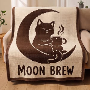 Op de afbeelding: Een crèmekleurige gehaakte deken met een donkerbruine rand en een grote donkerbruine halve maan afbeelding. Een donkerbruine kat zit op de maan en houdt een dampende mok vast. De tekst MOON BREW staat in donkerbruine blokletters eronder.