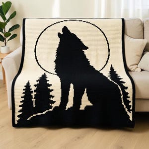 May include: A rectangular crochet blanket featuring a black silhouette of a wolf howling at a large, circular full moon against a cream-colored background. The design includes black pine tree silhouettes at the base of the wolf figure.