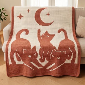 Op de afbeelding: Een rechthoekige gehaakte deken met een crèmekleurige achtergrond en een terracotta rand. Het ontwerp toont drie katten in terracotta, waarbij één kat naar een maansikkel en twee sterren erboven reikt.