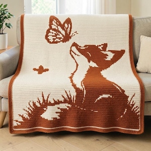 Puede incluir: Una manta de ganchillo rectangular con un diseño bicolor en naranja óxido y crema. El patrón muestra a un zorro sentado en la hierba mirando hacia una mariposa. La manta tiene un borde naranja óxido que enmarca el centro de color crema.