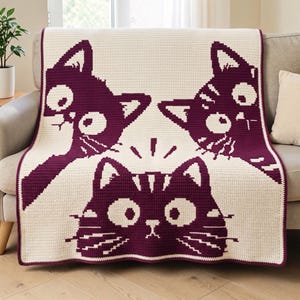Op de afbeelding: Een rechthoekige gehaakte deken met een crèmekleurige achtergrond en drie donkerpaarse kattengezichten. Twee katten bevinden zich in de bovenhoeken en één kat is onderaan gecentreerd, allemaal met grote ogen en snorharen.
