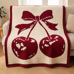 Puede incluir: Una manta de ganchillo rectangular con fondo color crema y un gran motivo de cerezas de color rojo oscuro centrado en la parte delantera. El diseño incluye dos cerezas conectadas por tallos a un lazo prominente, todo enmarcado por un borde rojo oscuro.
