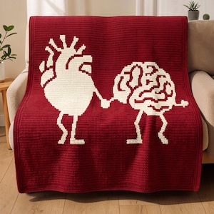 Könnte beinhalten: Eine tiefrote Häkeldecke mit einem großen, weißen, stilisierten anatomischen Herzen und einem weißen menschlichen Gehirn, beide mit dünnen, stabartigen Beinen dargestellt. Herz und Gehirn sind so positioniert, als würden sie sich in der Mitte halten.