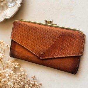 May include: A rectangular brown leather wallet featuring a diagonal embossed line pattern on the front flap and a gold-toned metal kiss-lock closure at the top. The wallet has a weathered texture and a pointed flap design that covers the main body.