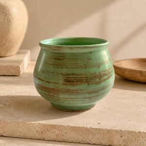 May include: A rounded ceramic pot featuring a light green glaze with horizontal, textured brown streaks across the exterior surface. The vessel has a wide, open top with a smooth, slightly flared rim and a narrow base, showcasing a rustic finish.