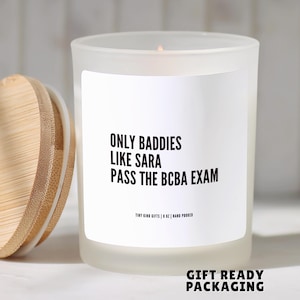 May include: A frosted glass candle in a jar with a bamboo lid. The white label features black text that reads: ONLY BADDIES LIKE SARA PASS THE BCBA EXAM. Below this, smaller text reads: TINY KIND GIFTS | 8 OZ | HAND POURED. The candle is lit.