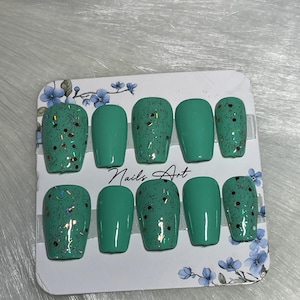 May include: A set of ten square-shaped, press-on nails. The nails are a solid teal colour, with some featuring a teal base and gold glitter and dark brown speckles. The card reads "Nails Art" in black script, with blue floral accents.