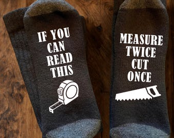 May include: A pair of dark grey socks featuring white text and graphics. The left sock displays the text IF YOU CAN READ THIS above a measuring tape icon. The right sock displays the text MEASURE TWICE CUT ONCE above a hand saw icon.