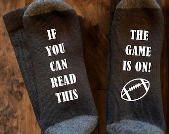 May include: A pair of dark grey crew socks with contrasting light grey heels and toes. The left sock features the white text IF YOU CAN READ THIS, and the right sock features the white text THE GAME IS ON! with a white graphic of a football.