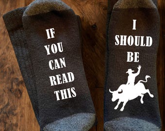 May include: A pair of dark grey socks with white text and graphics. The left sock features the text IF YOU CAN READ THIS in white capital letters. The right sock features the text I SHOULD BE above a white silhouette of a person riding a bucking bull.