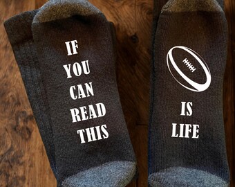 May include: A pair of dark grey crew socks with white text and a graphic. The left sock reads IF YOU CAN READ THIS in white capital letters. The right sock features a white graphic of an American football above the white text IS LIFE.