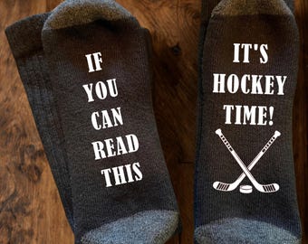 May include: A pair of dark grey socks with white text and graphics. The left sock reads IF YOU CAN READ THIS and the right sock reads IT'S HOCKEY TIME! with a graphic of two crossed hockey sticks and a puck positioned below the text on the right sock.
