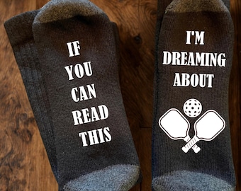 May include: A pair of dark grey crew socks with white text and graphics. The left sock reads IF YOU CAN READ THIS in white capital letters. The right sock reads I'M DREAMING ABOUT above a white graphic of two crossed pickleball paddles and a ball.