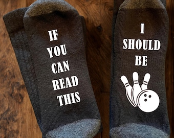 May include: A pair of dark grey socks with white text and graphics. The left sock features the text IF YOU CAN READ THIS in white. The right sock features the text I SHOULD BE and a white graphic of a bowling ball and three bowling pins.
