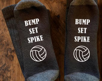 May include: A pair of dark charcoal grey crew socks featuring the white text BUMP SET SPIKE printed on the top of the foot, accompanied by a white graphic of a volleyball positioned directly below the text on each individual sock.